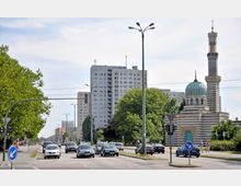 Straßenkreuzung in einer Stadt mit mehreren Fahrspuren und Autos, links Bäume und ein Gehweg, rechts ein auffälliges Gebäude mit grünlicher Kuppel und gestreiftem Minarett vor einem Hochhaus.