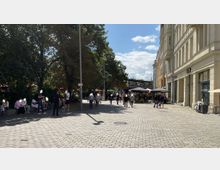 Eine gepflasterte Fußgängerzone mit Cafés und Geschäften auf der rechten Seite, darunter Starbucks, sowie Bäumen und Sitzbereichen auf der linken Seite. Einige Menschen sitzen oder spazieren in der sonnigen Umgebung.