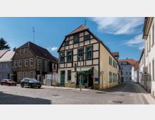 Fachwerkhaus an einer gepflasterten Straßenecke in einer Kleinstadt, umgeben von weiteren älteren Gebäuden mit Satteldächern. Verkehrsschilder und parkende Autos sind am Straßenrand sichtbar; im Hintergrund ein blauer Himmel mit wenigen Wolken.