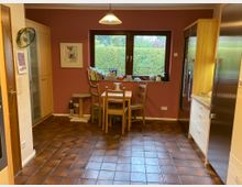 Eine Küche mit braunen Fliesenboden, roten Wänden und einer zentralen Fensterfront mit Blick auf eine grüne Hecke. Ein Holztisch mit vier Stühlen steht vor dem Fenster, umgeben von Holzschränken, einer Spiegelglasvitrine und Küchengeräten.