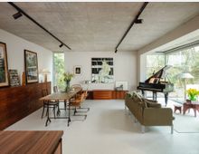 Open-plan modern living and dining room with a raw concrete ceiling, track lighting, and white walls. Mid-century wooden furniture, a dining table, sofa seating, and a grand piano sit beside a large floor-to-ceiling window looking out to a garden.