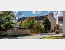 Wohngebiet mit gepflasterter Straße, mehrstöckigen Wohnhäusern mit roten Ziegeldächern und Schornsteinen. Im Vordergrund eine Steinmauer, ein Zaun mit Toren, Bäume und Büsche; rechts ein Gehweg und ein Stromkasten mit Graffiti.