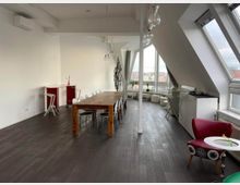 Bright loft-style meeting room with a long wooden table and chairs, white walls, and hanging pendant lights. Slanted ceiling with large skylight windows and a seating area by tall windows overlooking nearby rooftops.