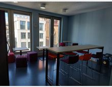 Heller Gemeinschaftsraum mit großen Fenstern, die auf moderne Gebäude und eine Backsteinkirche blicken. Der Raum ist mit hohen Tischen, bunten Stühlen und gepolsterten Würfeln möbliert, an der Wand hängen eine Uhr und Mülleimer stehen in einer Ecke.