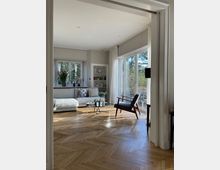 Wohnzimmer mit heller, moderner Einrichtung. Holzboden in Fischgrätmuster, eine weiße Couch mit Dekokissen, ein Glastisch und ein Sessel vor großen Fenstern mit weißen Vorhängen, die eine Aussicht auf Bäume bieten.