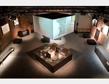 Industrieller Veranstaltungsraum mit Ziegelwänden, einer zentralen Bar in U-Form und hängendem, beleuchteten Würfel für Projektionen. Sitzgelegenheiten und Ausstellungstafeln sind entlang der Wände verteilt, die Beleuchtung erfolgt durch Deckenscheinwerfer.