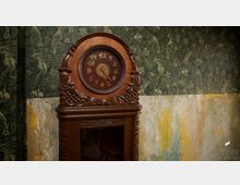 Holzstanduhr mit einem kunstvoll geschnitzten Gehäuse und römischen Zahlen auf dem Zifferblatt, vor einer Wand mit tropischem Muster und abblätternder Tapete darunter.