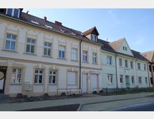 Mehrstöckige Reihenhäuser mit heller Fassade, teils beige, teils grün, und Giebeldächern. Vor den Gebäuden gibt es einen Gehweg mit frisch gepflastertem Bereich und einigen Blumentöpfen.