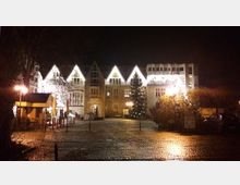 Nachtszene eines beleuchteten, historischen Gebäudes mit mehreren Satteldächern und großen Fenstern, flankiert von dekorativen Laternen. Vor dem Gebäude befinden sich ein gepflasterter Platz, ein geschmückter Weihnachtsbaum und Parkmöglichkeiten.