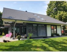 Modernes Einfamilienhaus mit Flachdachziegeln, großen Glasfenstern und Schiebetüren, umgeben von einem gepflegten Rasen und vereinzelten Pflanzen in Töpfen. Im Vordergrund ist eine Terrasse mit Sitzmöbeln und dekorativen Elementen zu sehen.