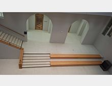 Innenansicht eines modernen Foyers mit hellen Steinböden, breiten Treppenstufen und Holzhandläufen sowie langen, niedrigen Holzpodesten. An der Rückwand befinden sich große Rundbögen und eine Holztür mit der Beschriftung „Museum“, daneben Fenster an der rechten Wand.