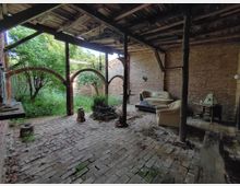 Alter Innenhof mit Ziegelboden und überdachtem Holzgebälk, bestehend aus alten Backsteinwänden, offenen Rundbögen mit Blick ins Grüne und vereinzelten Möbelstücken wie einem Sofa und einem Beistelltisch. Im Zentrum befindet sich eine kleine Pflanze, die umgeben von alten Gegenständen steht.