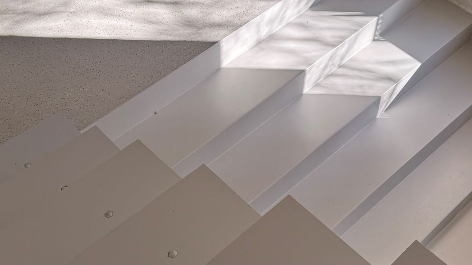 Interior view of a modern space with a wide white staircase leading up to a speckled terrazzo-like floor. Sunlight casts sharp rectangular shadows across the steps and landing near a white wall.