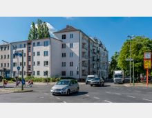 Straßenszene mit einem mehrstöckigen Wohnhaus im typischen Nachkriegsstil auf der linken Seite und einem älteren, detaillierter gestalteten Gebäude mit Balkonen rechts daneben. Im Vordergrund fahren Autos und ein LKW über eine Kreuzung, während Fußgänger, darunter Schulkinder, die Straße überqueren.