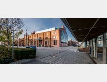 Backsteinbau mit großen, bogenförmigen Fenstern und modernen Glaselementen, umgeben von einem gepflasterten Platz und einzelnen Bäumen. Im Vordergrund ein Schild mit Straßenname „Hochschulring“ und moderne Überdachungen an der rechten Seite.