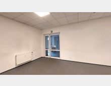 Ein leerer Raum mit weißen Wänden, grauem Bodenbelag und einer Decke mit Rasterplatten. An der Wand befindet sich ein Heizkörper, und auf der gegenüberliegenden Seite steht eine Glastür mit einem Türrahmen aus Metall, die zu einem gefliesten Bereich führt.