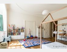 Helles Kinderzimmer mit Holzboden und einer offenen Raumgestaltung. Zu sehen sind ein Bücherregal, ein kleines Holzbettgestell in Hausform, ein farbenfroher Teppich, sowie ein Tisch mit Stühlen und Spielzeug.