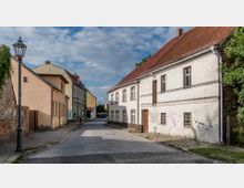 Schmale Kopfsteinpflasterstraße mit historischen Häusern auf beiden Seiten, darunter ein weißes Gebäude mit graffitibeschmierten Wänden und ein hellgelbes Gebäude. Im Hintergrund sind weitere Gebäude und Bäume unter einem teils bewölkten Himmel zu sehen.