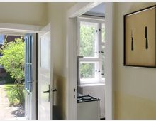 Ein heller Innenraum mit Blick durch eine geöffnete Haustür ins Freie, wo ein grüner Strauch sichtbar ist. Rechts im Bild hängt ein gerahmtes Bild mit einer Gabel und einem Messer, und im Hintergrund ist ein kleiner Bereich einer Küche mit einem Herd sowie ein Fenster mit geöffneten Fensterläden zu erkennen.