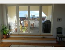 Blick aus einem hellen Wohnraum durch eine weiße Terrassentür auf einen Balkon mit Holzboden. Auf dem Balkon stehen ein Holztisch und Stühle, umgeben von Pflanzen und einer Dachlandschaft im Hintergrund.