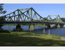Grüne Stahlbrücke mit geschwungenen Bögen (Glienicker Brücke) über einem Fluss, mit Parkbänken und einer Wiese im Vordergrund sowie Bäumen und klarem Himmel im Hintergrund.