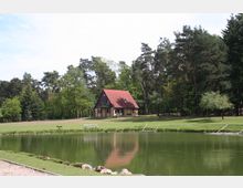 Ein kleines Haus mit rotem Satteldach steht am Rande eines Waldes auf einer grünen Wiese, im Vordergrund ein ruhiger Teich mit Steinufer und Holzstegen. Der Bereich ist von hohen Bäumen umgeben, die eine natürliche Umgebung schaffen.
