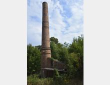 Hoher, runder Schornstein aus roten Ziegelsteinen neben einem kleinen, teilweise eingestürzten Backsteingebäude, umgeben von dichter Vegetation und Bäumen unter einem leicht bewölkten Himmel.