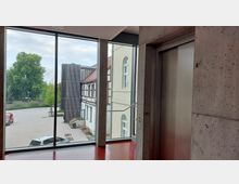 Blick durch eine große Glasfront auf einen Innenhof mit Pflastersteinen, umgeben von einem historischen Fachwerkgebäude und einem modernen Anbau. Rechts im Vordergrund befindet sich eine Metall-Aufzugstür in einer Betonwand.