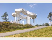 Ein moderner Aussichtsturm mit einer weißen, geometrischen Dachkonstruktion und schlanken Säulen, umgeben von einer Wiese mit bunten Wildblumen und einzelnen Bäumen. Im Vordergrund führt ein gepflasterter Weg den Hügel hinauf, unter einem klaren blauen Himmel.