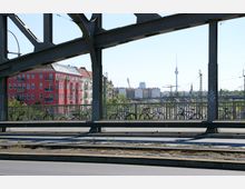 Städtische Szene aus Berlin, aufgenommen von einer Brücke mit stählernen Trägern; im Vordergrund Geländer mit abgestellten Fahrrädern, im Hintergrund ein rotes Wohngebäude und der Fernsehturm. Gleise und Stromleitungen verlaufen durch die untere Bildhälfte.