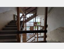 Treppenhaus in einem unvollendeten oder renovierungsbedürftigen Gebäude mit sichtbar rohen Holzgeländern und großen Fenstern, die nach außen auf eine Kiesfläche und Vegetation blicken. Die Wände sind weiß und teilweise beschädigt.