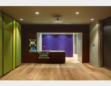 Moderne Küche mit dunklen Holzfronten, eingebauten Geräten und einer Arbeitsfläche mit Spüle. Im Hintergrund eine Wand in kräftigem Lila sowie ein Boden aus hellem Holz und eine dezente Deckenbeleuchtung.