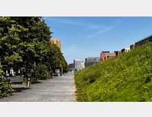 Städtische Szene mit einem gepflasterten Weg, flankiert von einer Baumreihe links und einer begrünten Böschung rechts. Im Hintergrund sind moderne Bürogebäude aus Glas und Backstein zu sehen, unter einem klaren, blauen Himmel.
