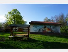 Eine grüne Wiese mit einer überdachten Holzbank im Vordergrund. Im Hintergrund befindet sich ein Gebäude mit einem bemalten Wandbild, das die Feuerwehr Vieraden darstellt, sowie umgebende Bäume und blauer Himmel.
