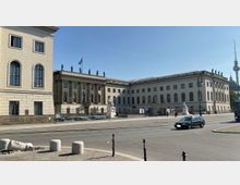 Straßenansicht der Humboldt-Universität zu Berlin mit klassizistischer Fassade, Säulenreihe und Statuen auf dem Dach. Im Vordergrund sind eine belebte Straße, Gehwege und ein Denkmal sichtbar, im Hintergrund ragt der Berliner Fernsehturm in den Himmel.