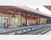 Ein Bahnsteig einer erhöhten U-Bahn-Station mit einem Dach aus Holzbalken und großen, bogenförmigen Fenstern. Im Vordergrund sind Sitzbänke, eine analoge Uhr, Fahrkartenautomaten und Hinweisschilder zu sehen; der Stationsname ist "Schlesisches Tor".