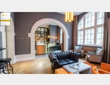 Lounge mit Polstermöbeln, Holzboden und großen Fenstern, die viel Tageslicht einlassen. Im Hintergrund befindet sich eine elegante Bar mit einer Holztheke, Barhockern und Flaschenregalen, eingerahmt von einem großen weißen Rundbogen.