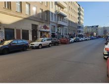 Straßenansicht in einer städtischen Umgebung mit Reihen von geparkten Autos auf beiden Seiten der Straße. Links sind mehrstöckige Altbauten mit Geschäften im Erdgeschoss zu sehen, während rechts modernere Gebäude stehen; im Hintergrund mündet die Straße in einen Platz.