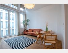 Helles Wohnzimmer mit großen Fenstern, die einen Blick auf ein modernes Gebäude bieten. Der Raum ist mit einem roten Sofa, einem runden Holztisch mit Stühlen, einer grünen Zimmerpflanze und einem gestreiften Teppich eingerichtet.