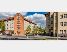 Straßenansicht mit Wohnhäusern aus der frühen Moderne, gekennzeichnet durch rote Ziegel- und beigefarbene Fassaden. Im Vordergrund stehen geparkte Autos, einige Bäume sowie Straßenschilder und ein Gehweg unter einem teils bewölkten Himmel.