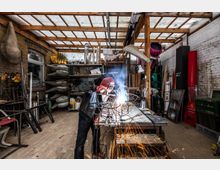 Ein Handwerker in einem Schweißhelm arbeitet an einer Metallkonstruktion, Funken fliegen von der Schweißstelle. Der Raum ist eine Werkstatt mit Ziegelwänden, Regalen voller Werkzeuge und Materialien sowie einer Holz- und Kunststoffdecke.