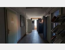 Ein schmaler Flur mit abgenutzten Wänden und gefliesten Böden in einem alten Gebäude, mit mehreren offenen Türen auf beiden Seiten. Links befindet sich eine beschriftete Zimmertür, rechts ein Regal mit Prospekten und ein Sicherheitsgitter am Fenster am Ende des Flurs, durch das Tageslicht einfällt.