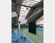 Indoor-Tennisplatz mit blauen und grünen Spielflächen, hohen Decken mit Oberlichtern und Metallverstrebungen. Im Vordergrund befinden sich Beleuchtungsausrüstung und ein Vorhang, der den Platz teilweise abtrennt.