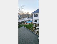 Ein moderner Gartenbereich mit einem rechteckigen Swimmingpool, umgeben von Rasen und gepflasterten Bereichen. Daneben steht ein helles Wohnhaus mit weißen Wänden und einem dunkelblauen Ziegeldach; im Hintergrund sind Bäume und eine Gartenmauer zu sehen.