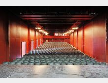 Ein leerer Theatersaal mit roten Wänden, einer schwarzen Decke und mehreren Reihen grüner Stühle, aufgenommen von der Bühne aus. Die Bühne wirkt abgenutzt, und im Hintergrund sind geschlossene Türen sowie ein kleiner Balkonbereich sichtbar.