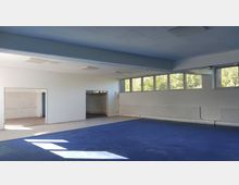 Ein leerer Raum mit hellen Wänden und blauer Bodenfläche. Große Fenster an der Oberseite der Wand lassen Tageslicht herein, und eine Türöffnung führt in einen angrenzenden Bereich mit weißen Fliesen.