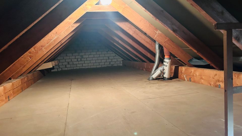 Ausgebauter Dachboden mit schrägen Dachflächen und freiliegenden Holzbalken, beleuchtet durch ein Dachfenster. Der Boden ist mit Platten ausgelegt; an der rechten Seite sind Lüftungsrohre und eine niedrige Abstellfläche zu sehen, im Hintergrund eine Ziegelwand.