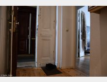 Blick in einen Hausflur mit geöffneter weißer Wohnungstür, die auf einen hölzernen Fußboden führt. Im Hintergrund sind eine dunkle Eingangstür sowie ein angrenzender Raum mit Sofa und Pelzdecke zu sehen.