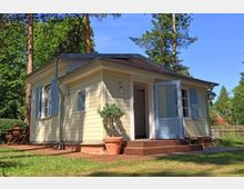 Kleines, einstöckiges Haus mit hellgelber Holzfassade, blauen Fensterläden und einer offenen Eingangstür, umgeben von hohen Bäumen und einem Garten mit Terrasse und Gartenmöbeln.
