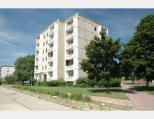 Mehrstöckiges Wohngebäude aus Betonplatten mit Balkonen, umgeben von Bäumen, Sträuchern und einer Grünfläche. Im Vordergrund führen ein Gehweg und eine schmale Straße durch die Szene.